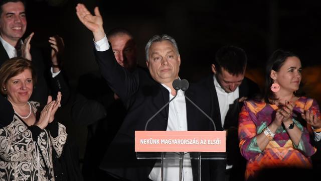 Orban steht im Dunkeln in Freien hinter zwei Mikrofonen und winkte mit erhobener Hand. Daneben zwei Frauen und drei Männer.