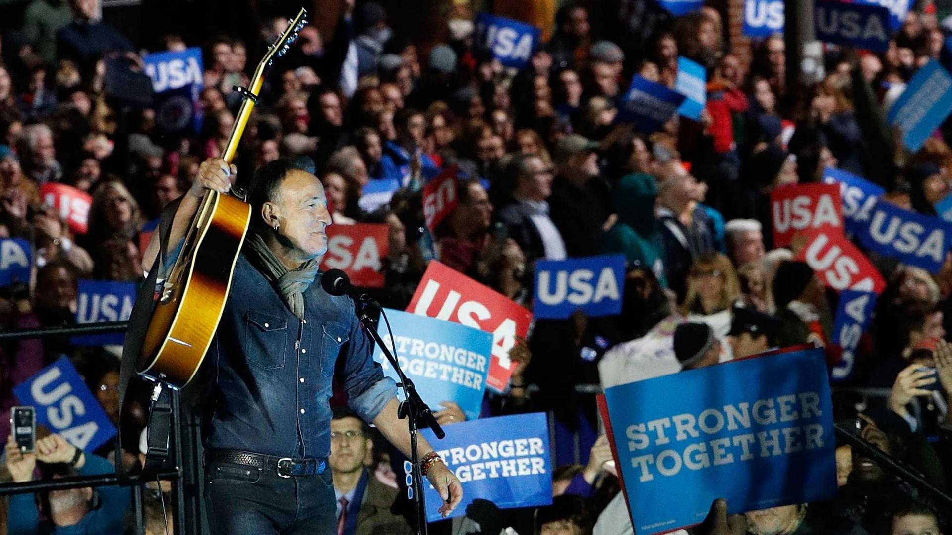Bruce Springsteen tritt vor den Wählern von Hillary Clinton auf und hält seine Gitarre zur Begrüßung in die Höhe. (2016) Bruce Springsteen tritt vor den Wählern von Hillary Clinton auf und hält seine Gitarre zur Begrüßung in die Höhe. (2016)