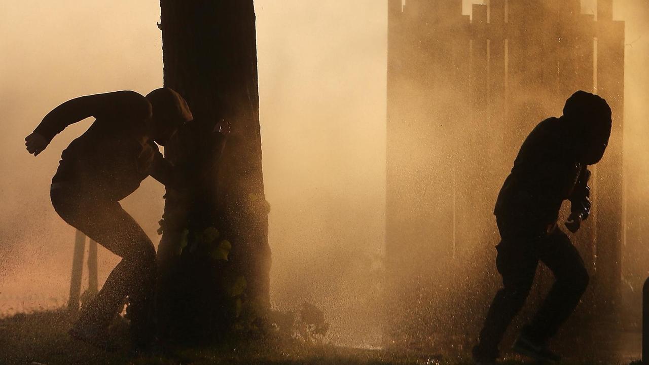 Die zwei Aktivisten, der Baum und die Barrikade sind als Silhouetten vor dem von Wasserspritzern durchsetzten Gegenlicht zu sehen. Die zwei Aktivisten, der Baum und die Barrikade sind als Silhouetten vor dem von Wasserspritzern durchsetzten Gegenlicht zu sehen.