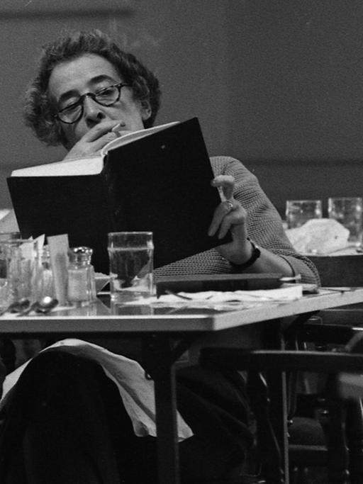 Die Philosophin Hannah Arendt sitzt an einem Tisch in einer Bibliothek, liest und raucht.