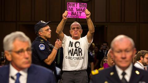 Chuck Hagel und Martin Dempsey sitzen an einem Tisch, während hinter ihnen der Aktivist Tighe Berry aufsteht. Dieser trägt ein T-Shirt mit der Aufschrift "No War" und ein pinkfarbenes Schild mit dem Text "Don´t take Isil war bait"