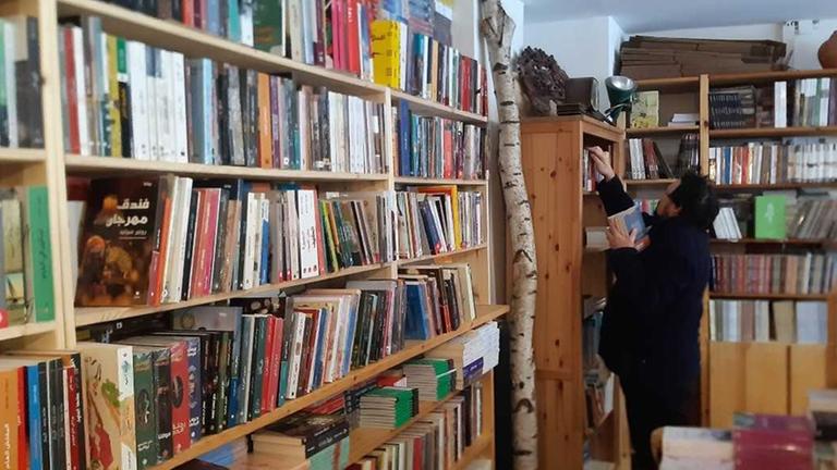 Zu sehen sind Regale voller Bücher. In dem Raum ist auch ein Birkenstamm als Dekoration aufgestellt. Ein Mann sortiert Bücher in einen Schrank.