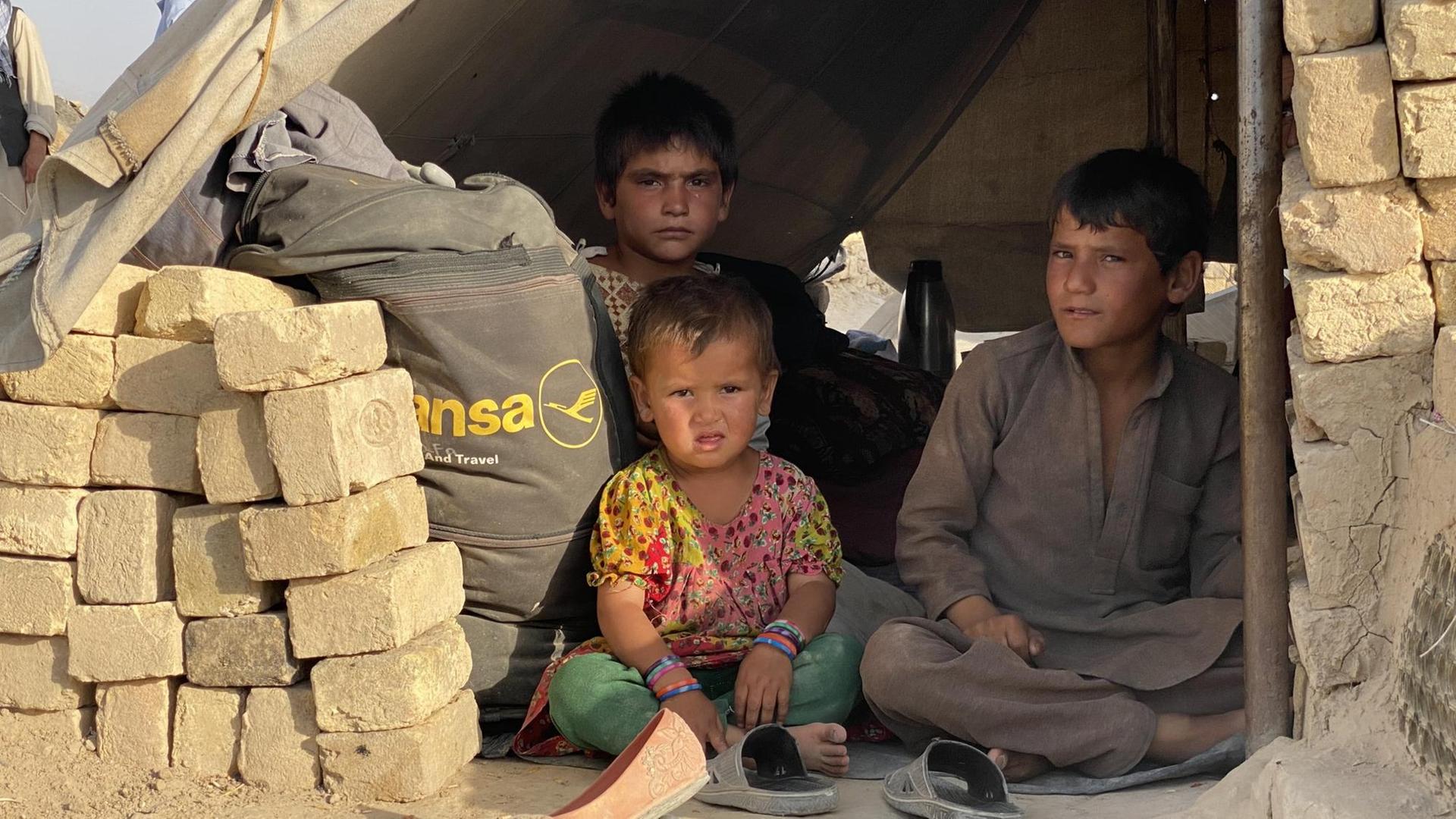 Drei Kinder sitzen in einer Kombination aus Steinbau und Zelt auf dem Boden.