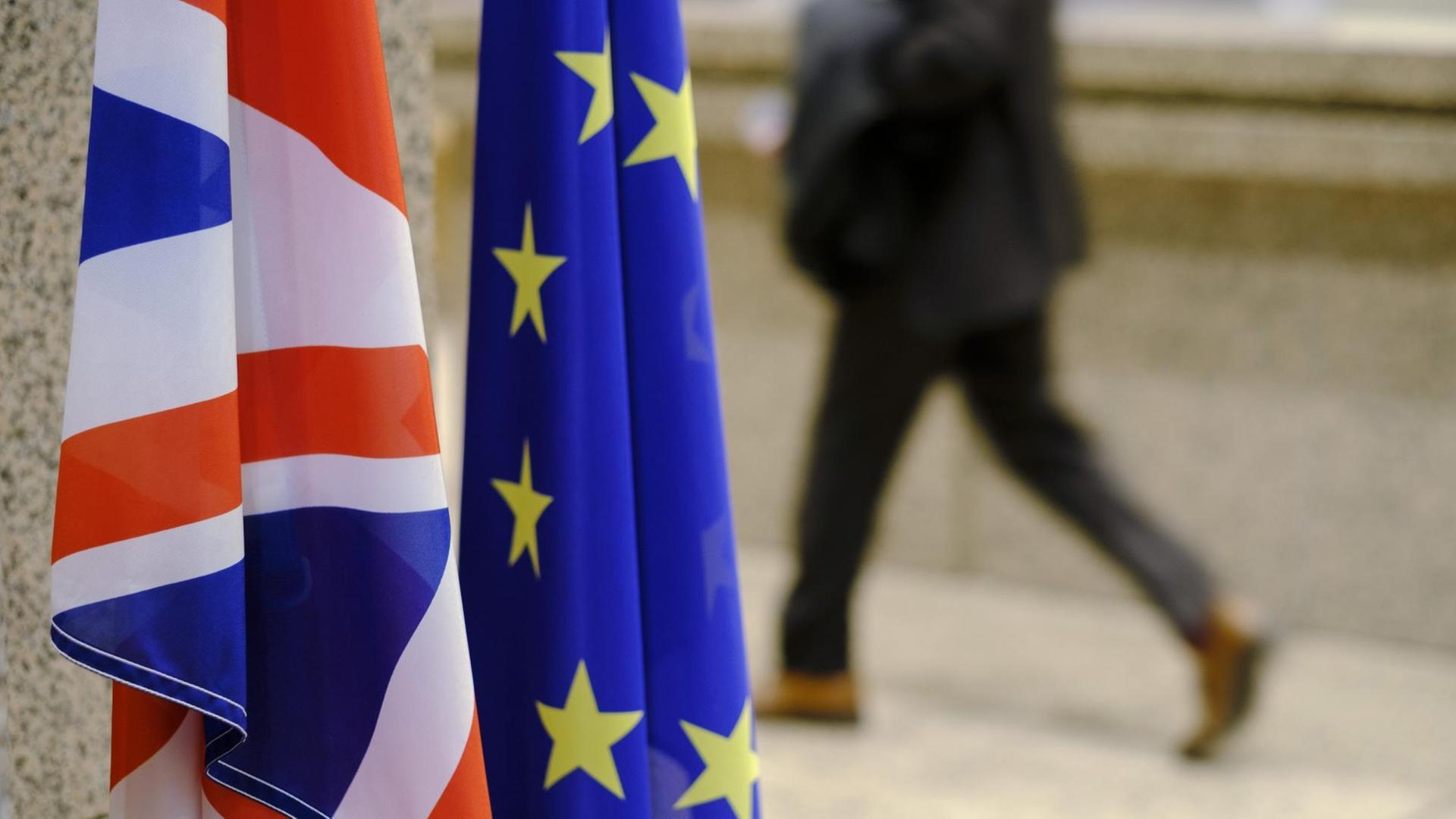 Die Fahnen Großbritanniens und der EU nebeneinander im Vordergrund, im Hintergrund ein laufender Mensch auf der Straße Die Fahnen Großbritanniens und der EU nebeneinander im Vordergrund, im Hintergrund ein laufender Mensch auf der Straße