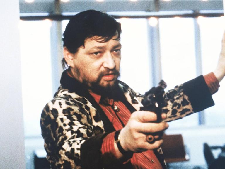 Rainer Werner Fassbinder als Schauspieler in einer Szene des Films "Kamikaze 1989". Er hat eine Pistole in der Hand und richtet sie offenbar auf jemand, der nicht zu sehen ist.