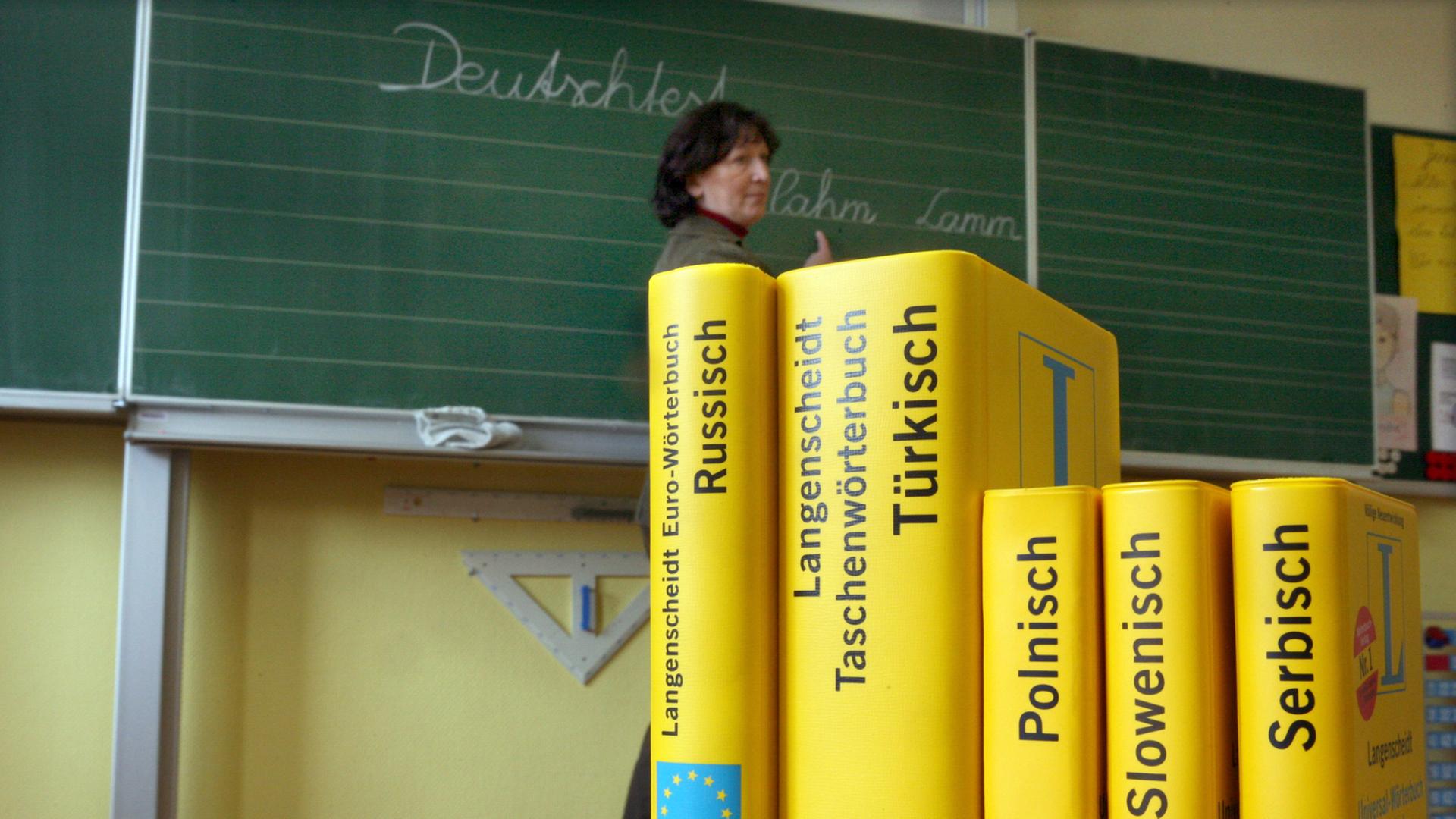 Mehrere Wörterbücher stehen auf einem Tisch in einer Schule. Im Hintergrund ist eine Tafel zu sehen, vor der eine Frau steht. Mehrere Wörterbücher stehen auf einem Tisch in einer Schule. Im Hintergrund ist eine Tafel zu sehen, vor der eine Frau steht.
