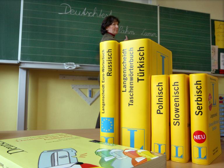Mehrere Wörterbücher stehen auf einem Tisch in einer Schule. Im Hintergrund ist eine Tafel zu sehen, vor der eine Frau steht.