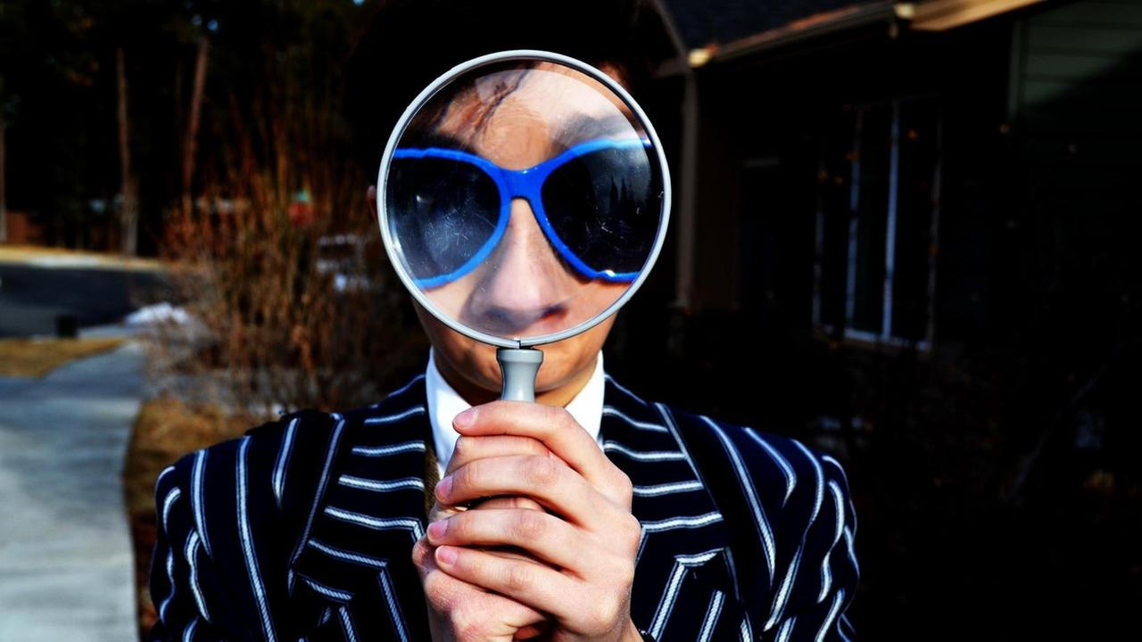 Eine Person mit Sonnenbrille hält eine Lupe vor dem Gesicht. Eine Person mit Sonnenbrille hält eine Lupe vor dem Gesicht.