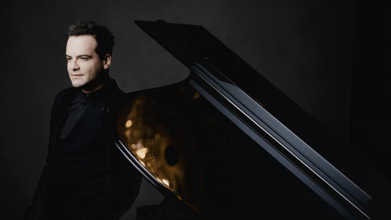 Der Pianist steht neben einem aufgeklappten Flügel, in dem sich goldene Lichter spiegeln. Der Pianist steht neben einem aufgeklappten Flügel, in dem sich goldene Lichter spiegeln.