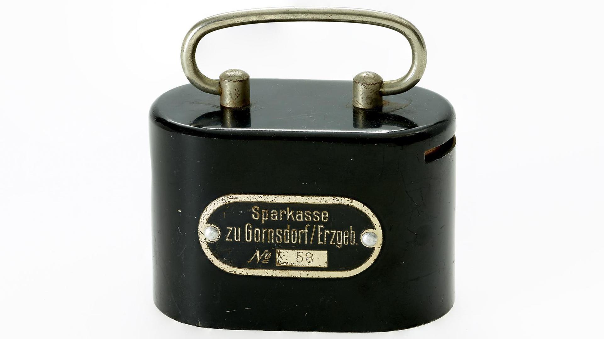 Heimsparbüchse Nr. 58 der Sparkasse Gornsdorf (Erzgebirge), 1912 - Eine schwarze, ovale Blechdose mit Henkel und Sparschlitz an der rechten Seite. Heimsparbüchse Nr. 58 der Sparkasse Gornsdorf (Erzgebirge), 1912 - Eine schwarze, ovale Blechdose mit Henkel und Sparschlitz an der rechten Seite.