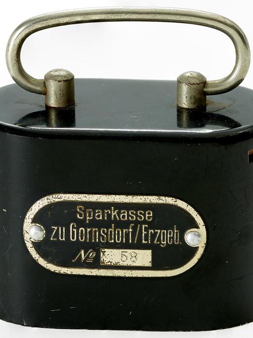 Heimsparbüchse Nr. 58 der Sparkasse Gornsdorf (Erzgebirge), 1912 - Eine schwarze, ovale Blechdose mit Henkel und Sparschlitz an der rechten Seite.