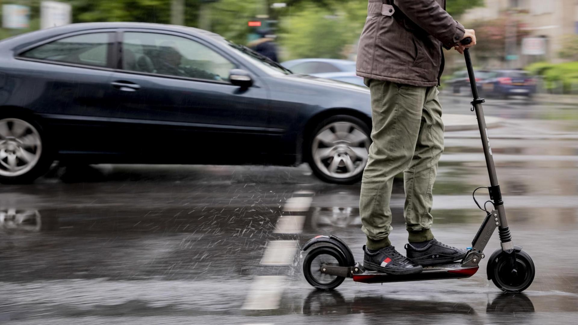 Auf einer nassen Strass fährt im Vordergund ein E-Scooter, im Hintergrund in der Unschärfe sind weitere Autos zu sehen. Auf einer nassen Strass fährt im Vordergund ein E-Scooter, im Hintergrund in der Unschärfe sind weitere Autos zu sehen.