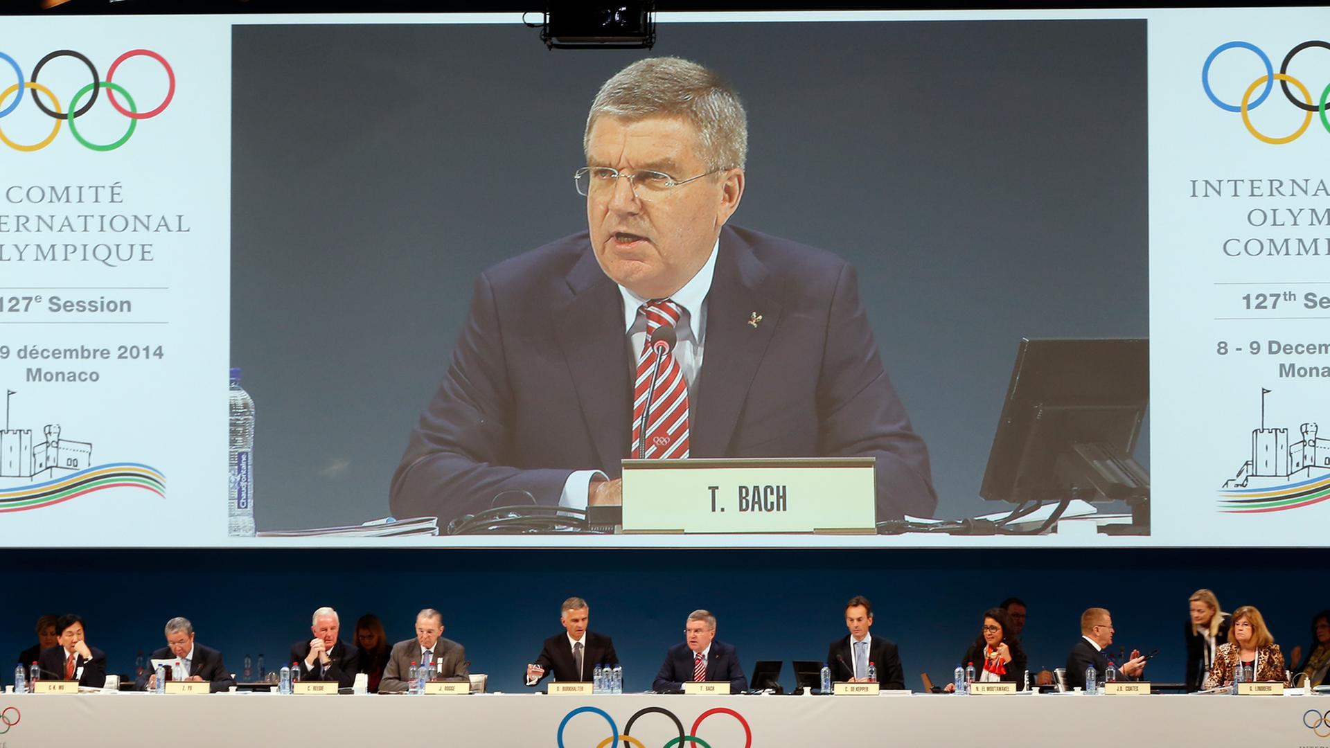 IOC-Präsident Bach sitzt neben den anderen Vorstandsmitgliedern und spricht vor dem Komittee beim Reformgipfel in Monaco. Über Bach selbst ist er noch einmal auf einem übergroßen Bildschirm zu sehen. IOC-Präsident Bach sitzt neben den anderen Vorstandsmitgliedern und spricht vor dem Komittee beim Reformgipfel in Monaco. Über Bach selbst ist er noch einmal auf einem übergroßen Bildschirm zu sehen.