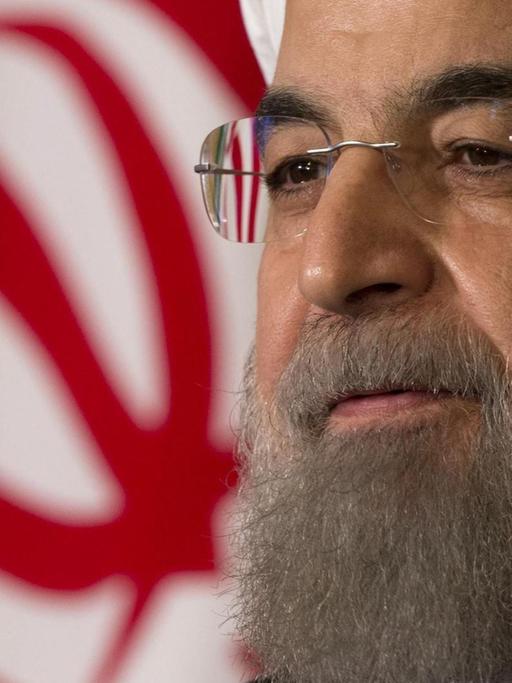Rohani trägt einen weißen Turban und Brille. Hinter ihm sieht man einen teil der iranischen Nationalflagge.