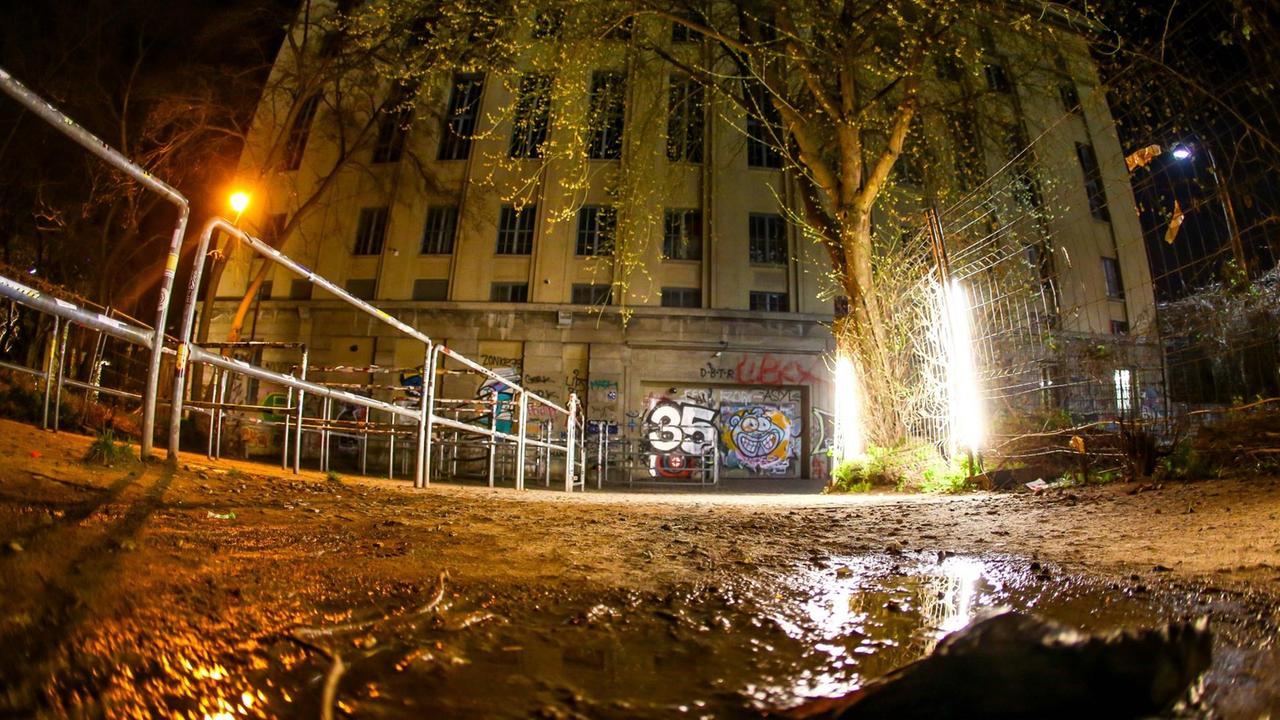Das Vorfeld des Berliner Clubs Berghain bei Nacht. Einige Lampen brennen und geben etwas Licht, Stahlgeländer sind zu erkennen, wo sich bei Betrieb die Gäste anstellen. Im Vordergründ ist eine Pfütze. Menschen sind nicht zu sehen.