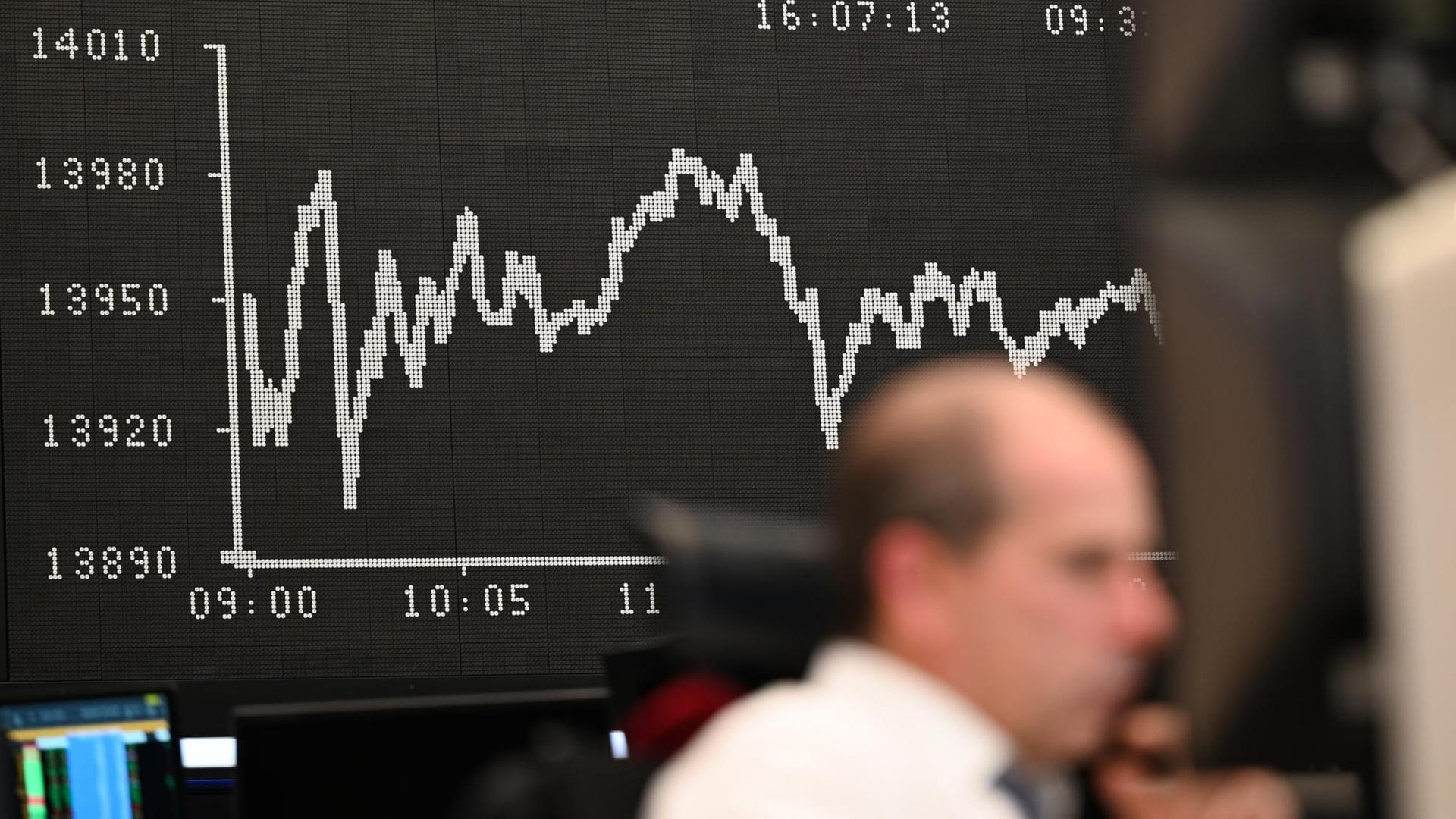 Impression von der Börse in Frankfurt: Während ein Aktienhändler an der Börse seine Monitore beobachtet, wird auf einer Tafel im Hintergrund die Dax-Kurve angezeigt.