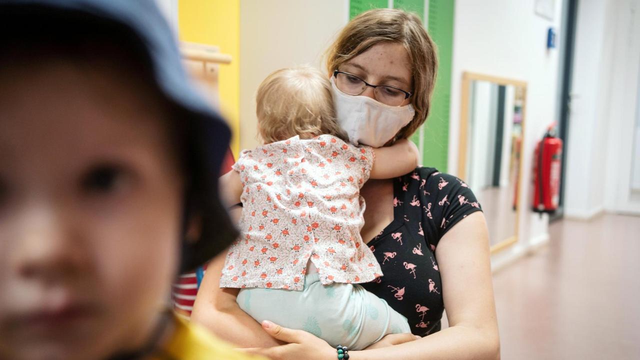 Eine Erzieherin mit Mundschutz, hält einen ihrer Schützlinge in einer Kindertagesstätte im Arm. Eine Erzieherin mit Mundschutz, hält einen ihrer Schützlinge in einer Kindertagesstätte im Arm.