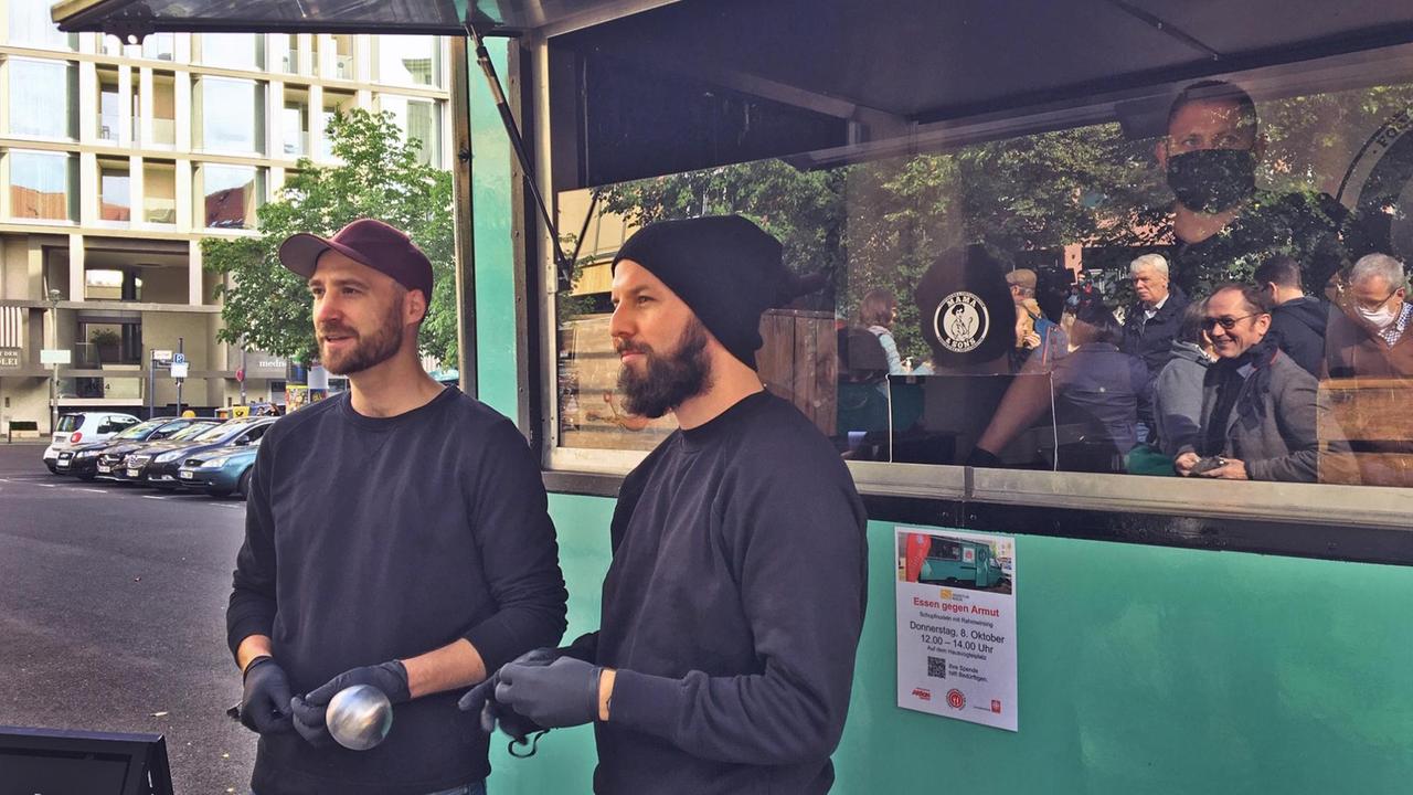André Johst und Janis Gensch, zwei Junge Männer mit Bart in Freizeitkleidung, vor ihrem grünen Imbisswagen, der als Foodtruck der Berliner Caritas im Einsatz ist