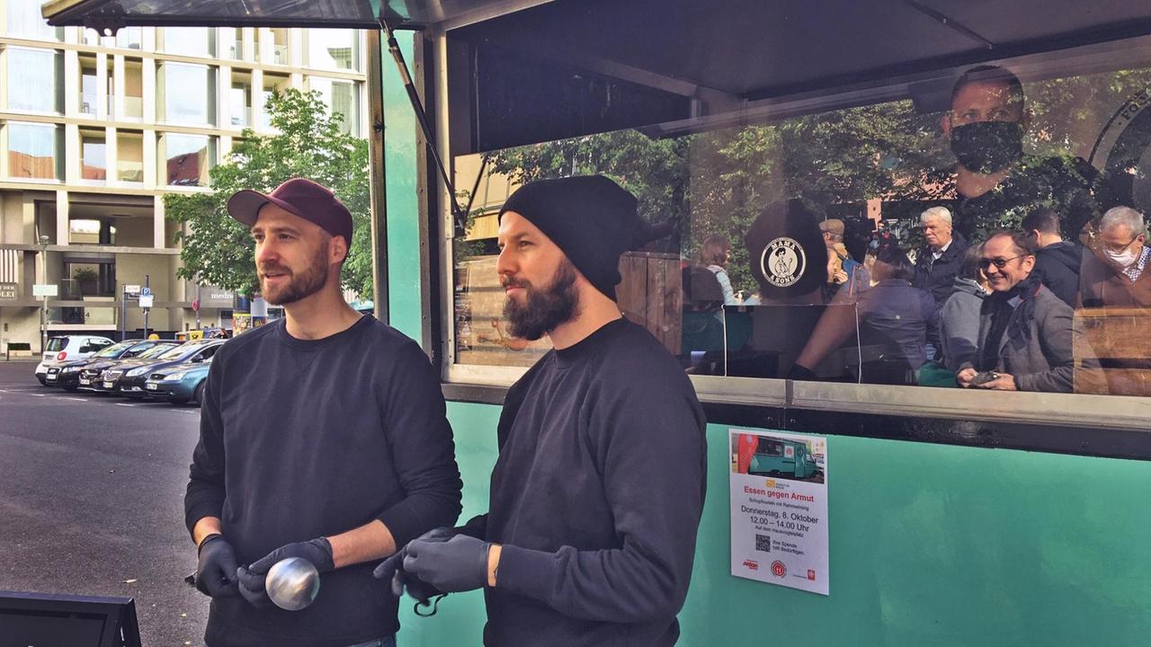 André Johst und Janis Gensch, zwei Junge Männer mit Bart in Freizeitkleidung, vor ihrem grünen Imbisswagen, der als Foodtruck der Berliner Caritas im Einsatz ist André Johst und Janis Gensch, zwei Junge Männer mit Bart in Freizeitkleidung, vor ihrem grünen Imbisswagen, der als Foodtruck der Berliner Caritas im Einsatz ist