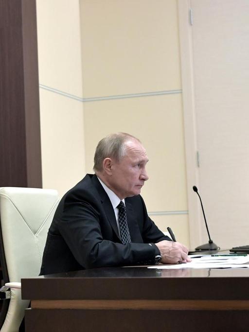 Wladimir Putin sitz an seinem Schreibtisch vor einem Computermonitor, auf dem ein Bild des Kremls zu sehen ist.