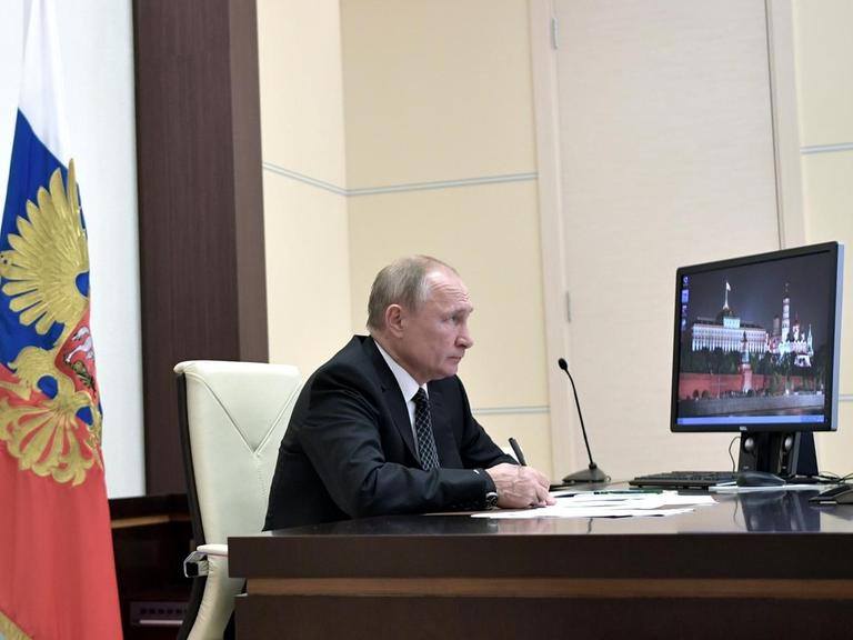 Wladimir Putin sitz an seinem Schreibtisch vor einem Computermonitor, auf dem ein Bild des Kremls zu sehen ist.