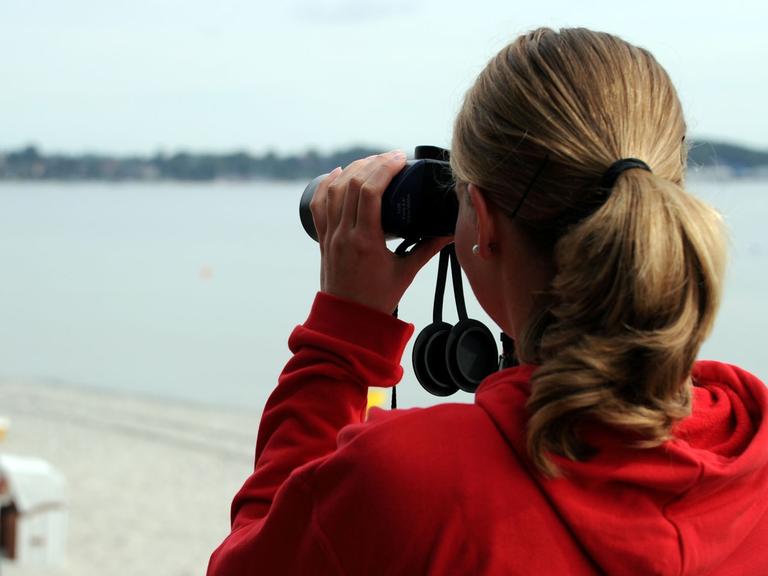 Ein freiwilliges Mitglied der Deutschen Lebensrettungsgesellschaft (DLRG) blickt am 19.08.2013 mit einem Fernglas von der Station auf den Strand von Eckernförde (Schleswig-Holstein).