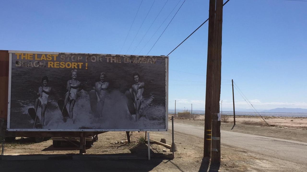 Ein Plakat am Rande einer Straße in trister Landschaft zeigt Jetskiing bei Bombay Beach. Ein Plakat am Rande einer Straße in trister Landschaft zeigt Jetskiing bei Bombay Beach.