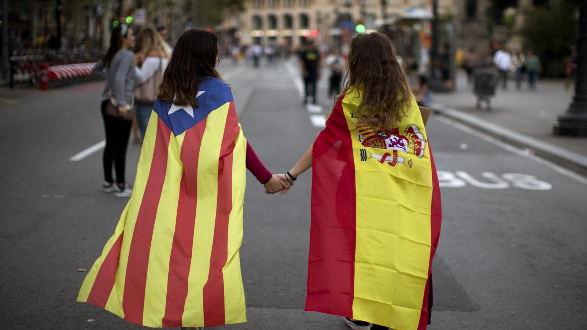 Zwei junge Frauen gehen nebeneinander. Ein von ihnen trägt eine Spanien-Flagge als Umhang, die andere eine Flagge von Katalonien. Zwei junge Frauen gehen nebeneinander. Ein von ihnen trägt eine Spanien-Flagge als Umhang, die andere eine Flagge von Katalonien.
