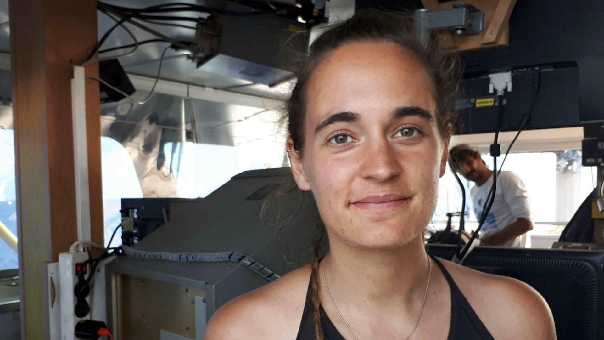 Kapitänin Carola Rackete steht auf der Brücke ihres Schiffes und lächelt in die Kamera. Dahinter unscharf ein Mann. Kapitänin Carola Rackete steht auf der Brücke ihres Schiffes und lächelt in die Kamera. Dahinter unscharf ein Mann.