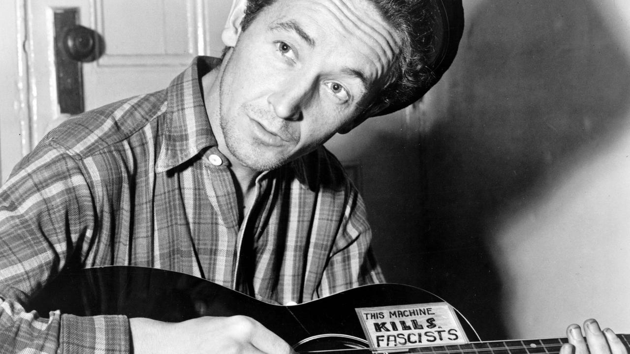 Ein Mann mit Mütze sitzt vor einer Tür und spielt auf einer Gitarre, die die Aufschrift "THIS MACHINE KILLS FASCISTS" zu sehen ist. Ein Mann mit Mütze sitzt vor einer Tür und spielt auf einer Gitarre, die die Aufschrift "THIS MACHINE KILLS FASCISTS" zu sehen ist.