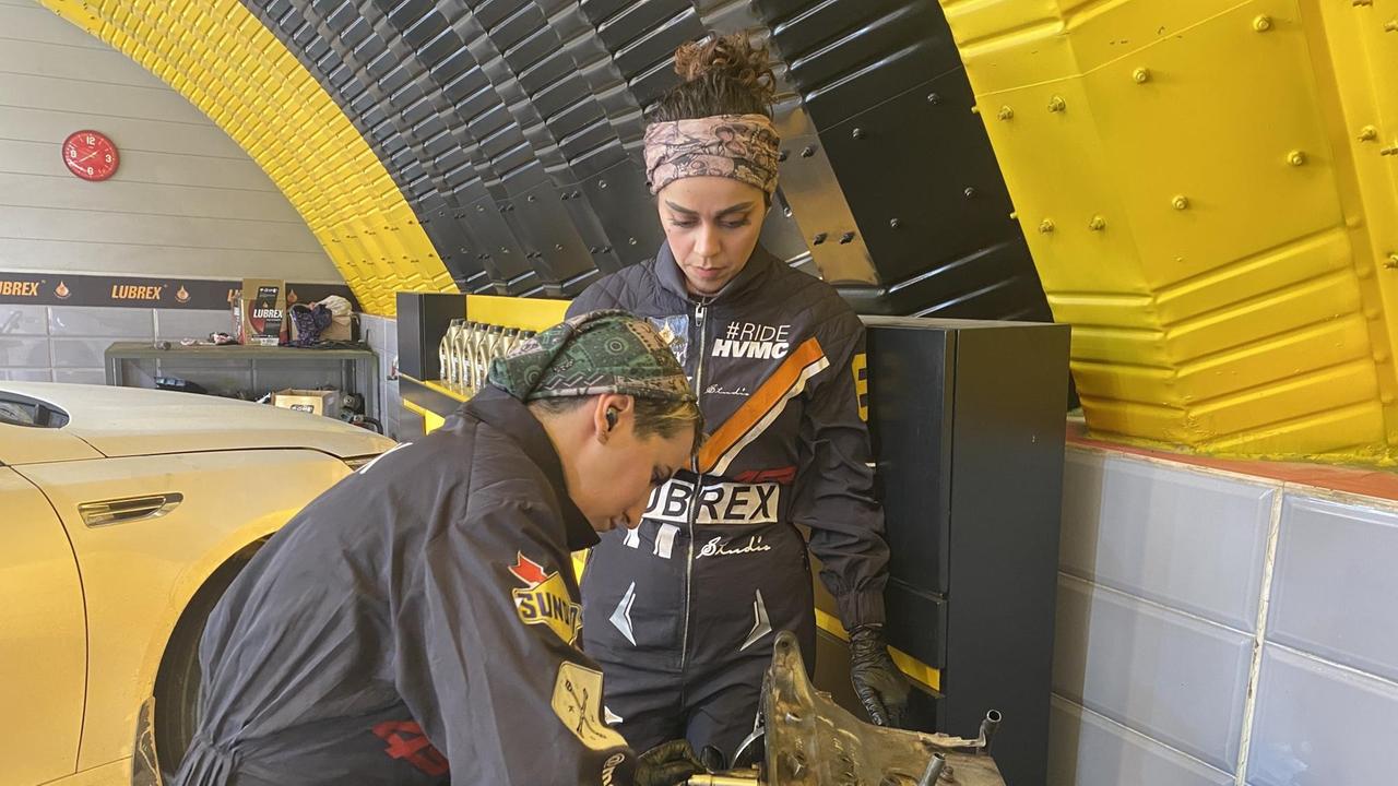 Zwei junge Frauen mit Haarband und schwarzen Overalls beugen sich über ein Autoteil. Zwei junge Frauen mit Haarband und schwarzen Overalls beugen sich über ein Autoteil.