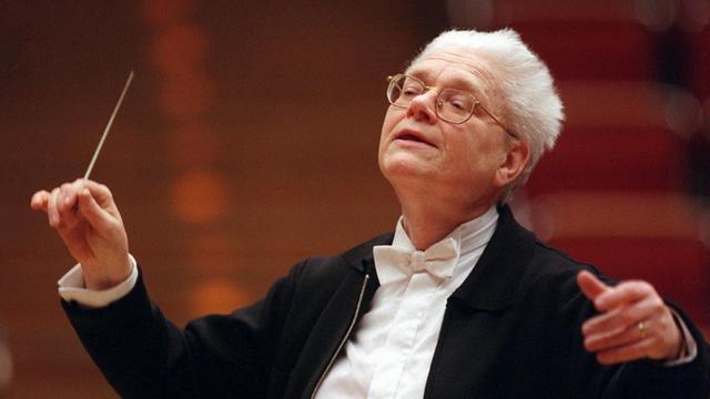 Der Dirigent Hans Zender dirigiert mit dem Taktstock in der rechten Hand, er trägt einen Frack mit weißem Hemd und weißer Fliege, er hat weißes, zurückgekämmtes Haar und trägt eine helle Brille.