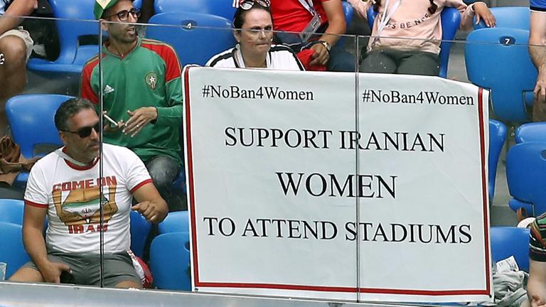 Das Bild zeigt eine Szene am Rande der Fußball-WM-Vorrundenbegegnung zwischen Marokko und dem Iran im Sankt-Petersburg-Stadion. Ein Poster zur Unterstützung von iranischen Frauen zum Besuch in Fußball-Stadien hängt an der Tribüne.