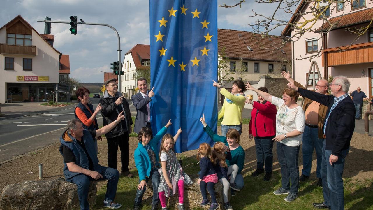 Auf einem Platz im Veithshöchheimer Ortsteil Gadheim steht eine Gruppe Erwachsene und Kinder und deutet auf eine gehisste Flagge der Europäischen Union. 