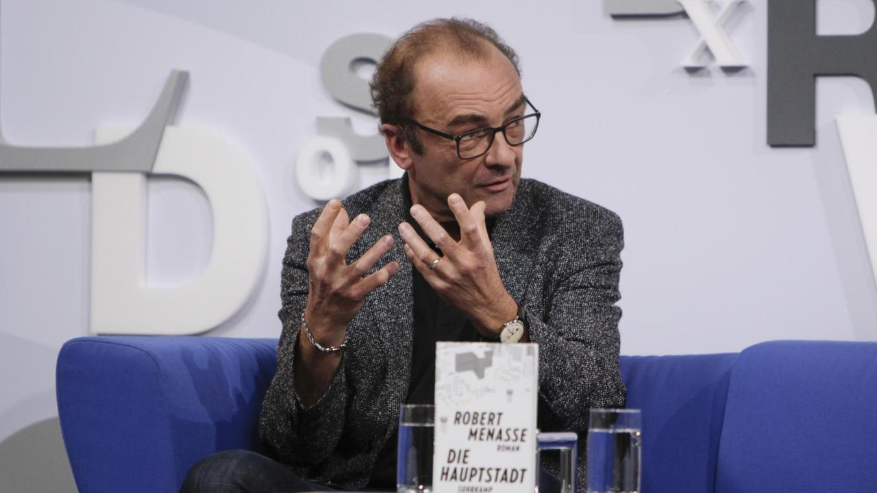 Der Schriftsteller Robert Menasse auf einem blauen Sofa, mit seinem Buch "Die Hauptstadt" auf dem Tisch, auf der Frankfurter Buchmesse. Der Schriftsteller Robert Menasse auf einem blauen Sofa, mit seinem Buch "Die Hauptstadt" auf dem Tisch, auf der Frankfurter Buchmesse.