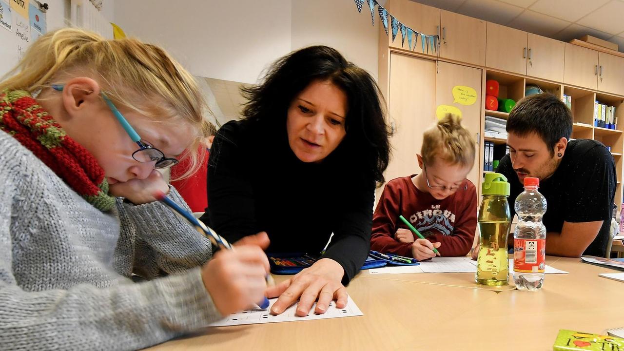 Eine Schulassistentin und ein Schulassistent sitzen mit am Gruppentisch in der Klasse und helfen den Kindern. Eine Schulassistentin und ein Schulassistent sitzen mit am Gruppentisch in der Klasse und helfen den Kindern.