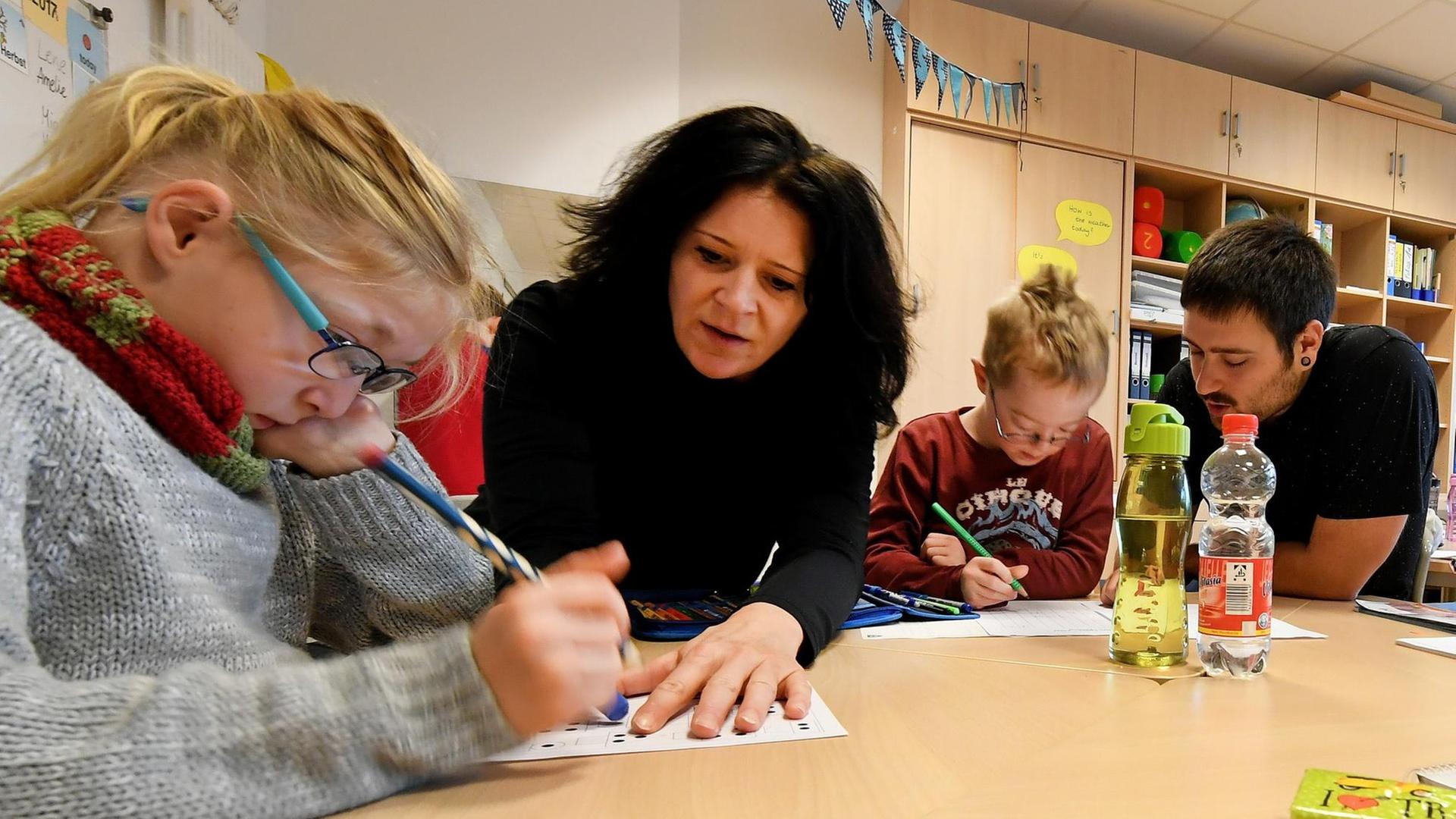 Eine Schulassistentin und ein Schulassistent sitzen mit am Gruppentisch in der Klasse und helfen den Kindern. Eine Schulassistentin und ein Schulassistent sitzen mit am Gruppentisch in der Klasse und helfen den Kindern.