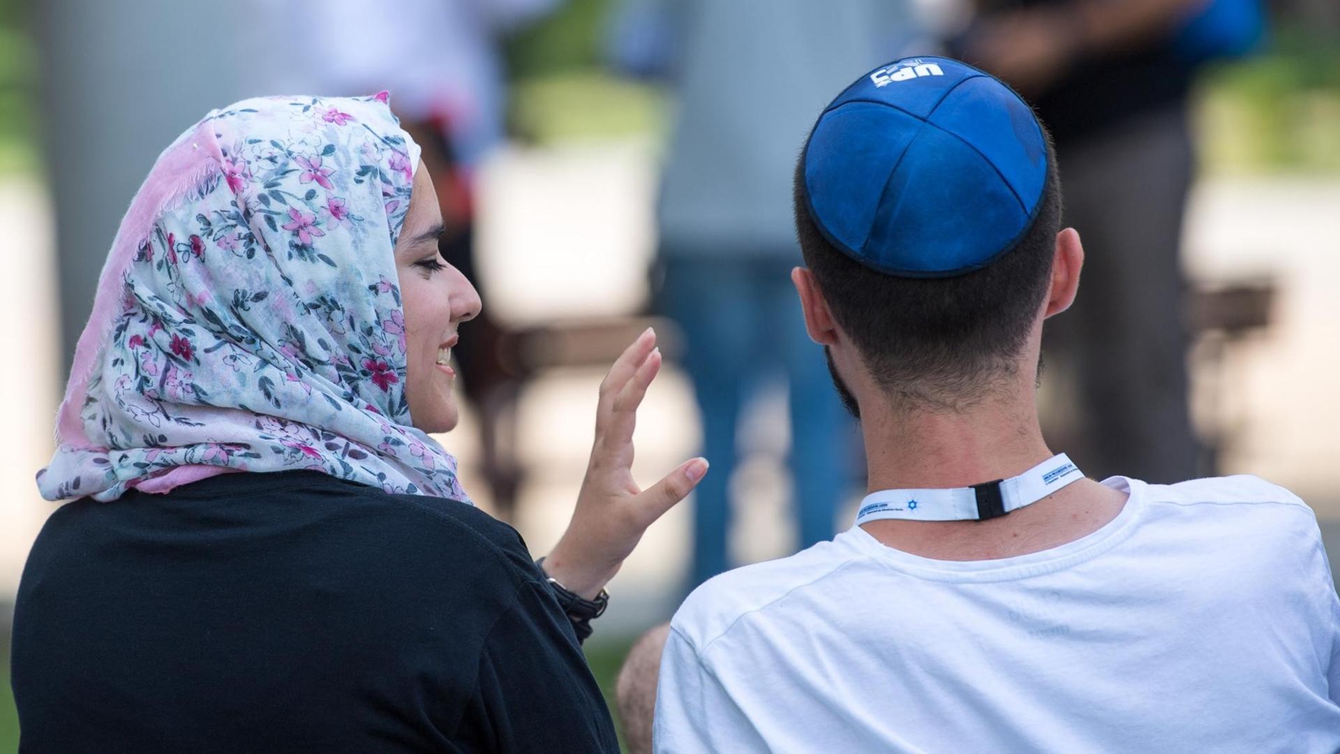Eine junge Muslima mit Kopftuch und ein junger jüdischer Mann mit Kippa unterhalten sich miteinander. Sie sitzen auf einer Bank, vom Betrachter abgewandt. Eine junge Muslima mit Kopftuch und ein junger jüdischer Mann mit Kippa unterhalten sich miteinander. Sie sitzen auf einer Bank, vom Betrachter abgewandt.