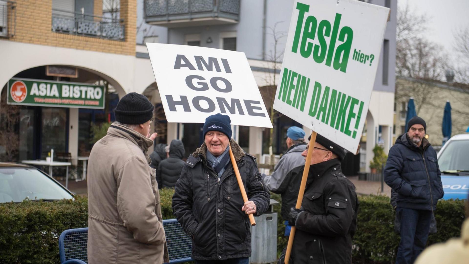 Auf der Straße stehen vier Männer, zwei von ihnen tragen Schilder, auf denen steht "Ami go home" und "Tesla nein danke"