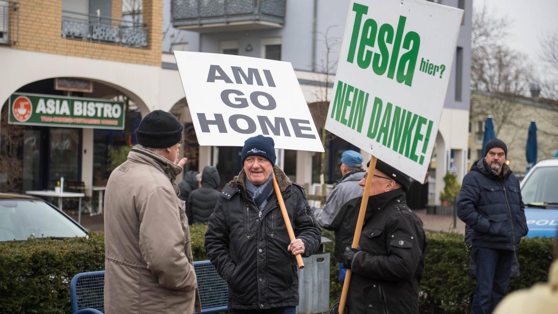 Auf der Straße stehen vier Männer, zwei von ihnen tragen Schilder, auf denen steht "Ami go home" und "Tesla nein danke" Auf der Straße stehen vier Männer, zwei von ihnen tragen Schilder, auf denen steht "Ami go home" und "Tesla nein danke"
