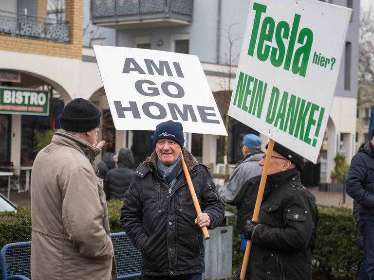 Auf der Straße stehen vier Männer, zwei von ihnen tragen Schilder, auf denen steht "Ami go home" und "Tesla nein danke"