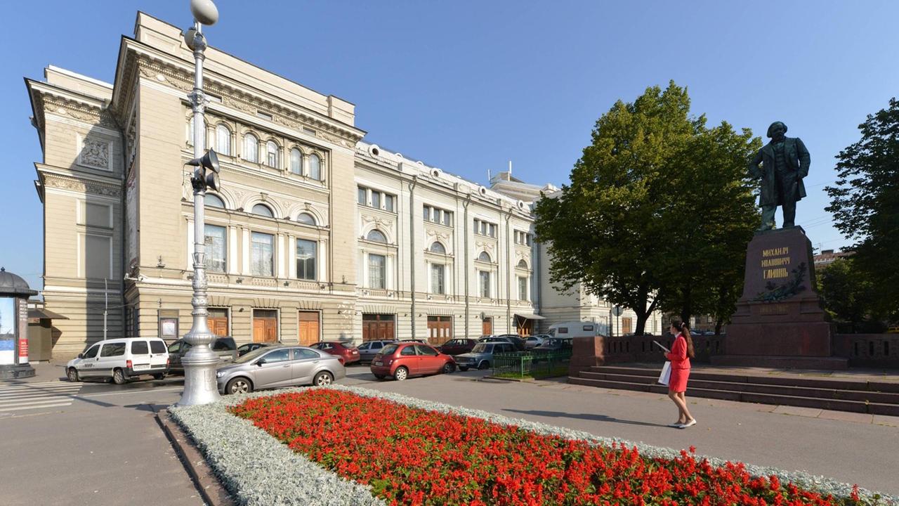 Blick über eine Blumenrabatte samt Denkmal eines Mannes zu einem historischen, großen Gebäude mit vielen Fenstern. Blick über eine Blumenrabatte samt Denkmal eines Mannes zu einem historischen, großen Gebäude mit vielen Fenstern.