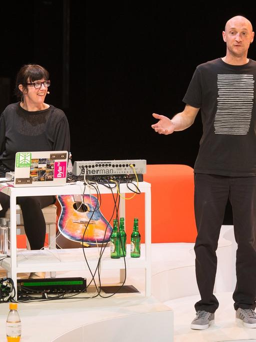 Ein Mann mit schwarzen T-Shirt steht auf einer weißen Bühne, hinter ihm sitzt eine Frau am Klavier.
