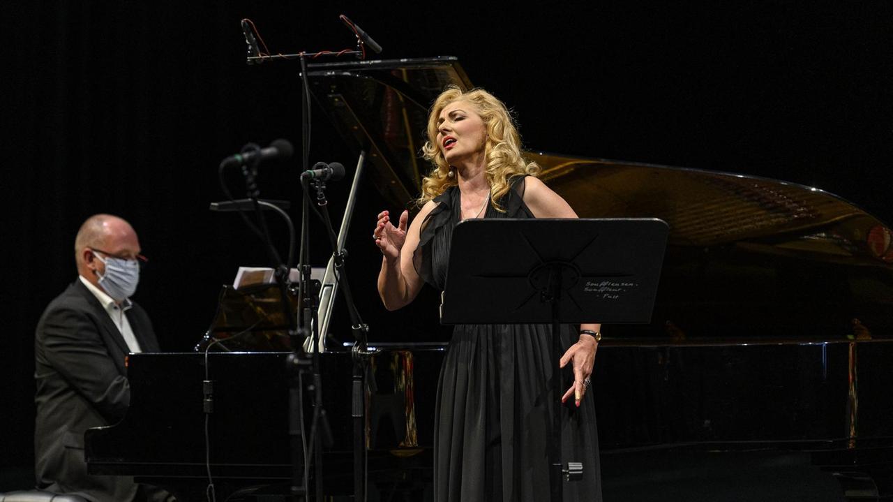 Eine blonde Sängerin in schwarzem Abendkleid steht neben einem Flügel samt Pianisten. Eine blonde Sängerin in schwarzem Abendkleid steht neben einem Flügel samt Pianisten.