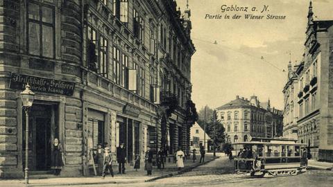 Historische Stadtansicht von Gablonz an der Neiße: Stadthäuser säumen eine Straße mit Kopfsteinpflaster, über die eine Straßenbahn fährt.