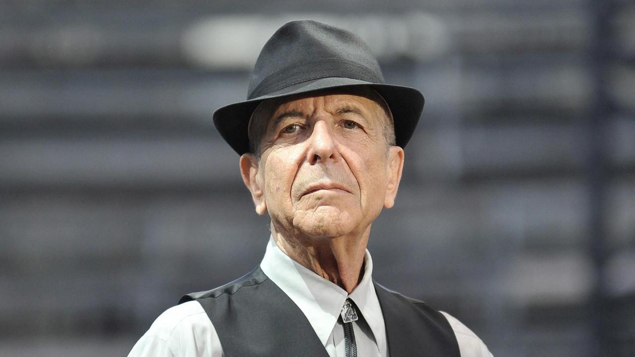 Leonard Cohen trägt ein helles Hemd, darüber eine dunkle Weste, außerdem einen Hut und schaut in die Kamera. Leonard Cohen trägt ein helles Hemd, darüber eine dunkle Weste, außerdem einen Hut und schaut in die Kamera.