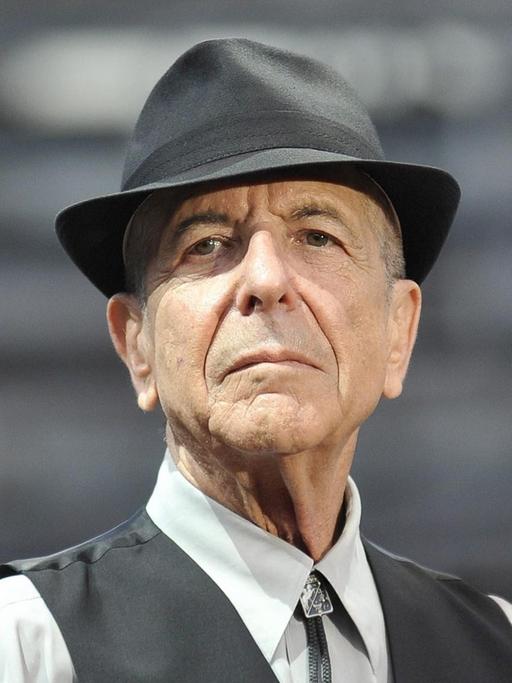 Leonard Cohen trägt ein helles Hemd, darüber eine dunkle Weste, außerdem einen Hut und schaut in die Kamera.