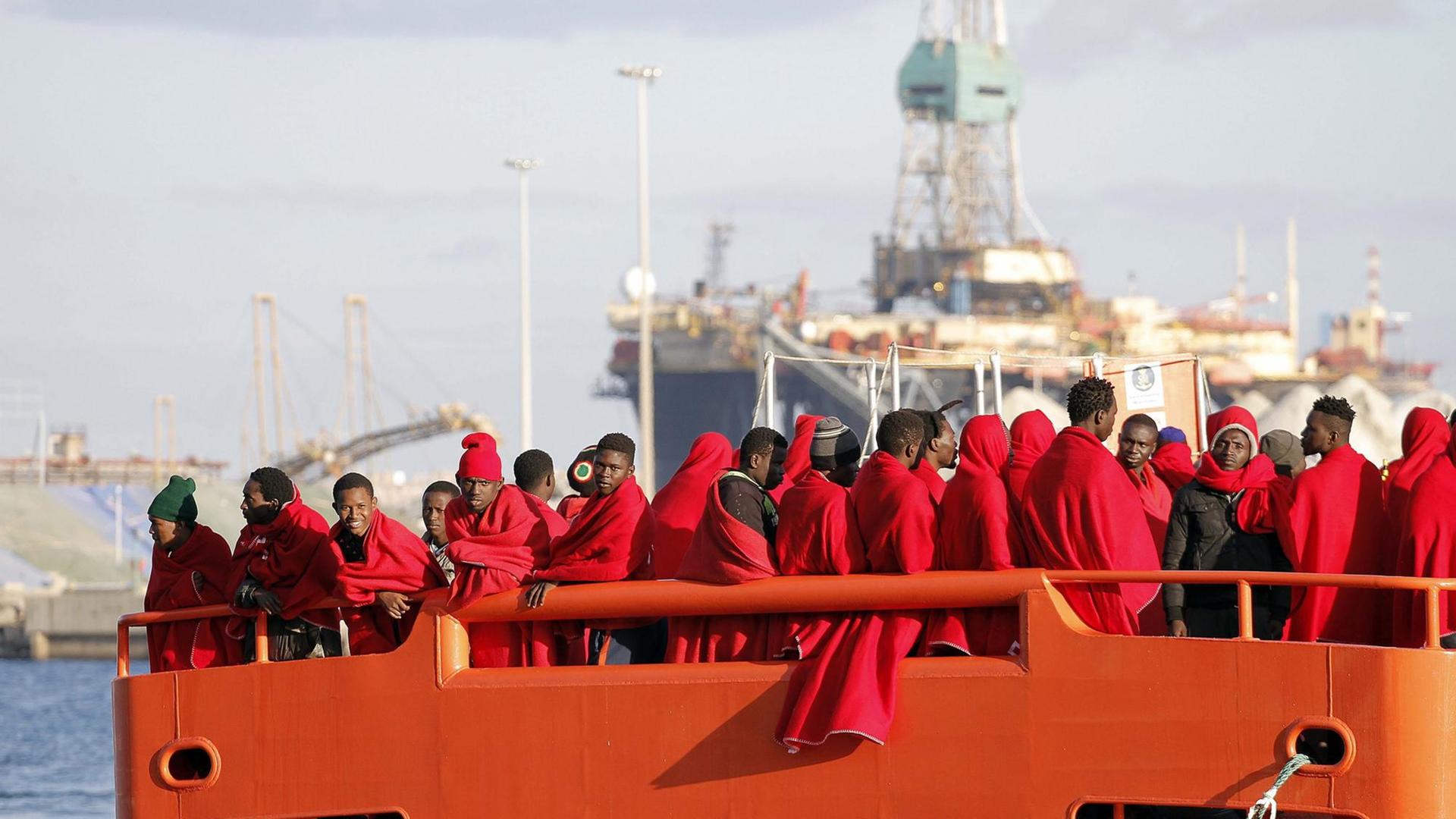 Gerettete Flüchtlinge aus der Subsahara stehen auf einem Boot der spanischen Küstenwache, eingewickelt in rote Decken. Gerettete Flüchtlinge aus der Subsahara stehen auf einem Boot der spanischen Küstenwache, eingewickelt in rote Decken.