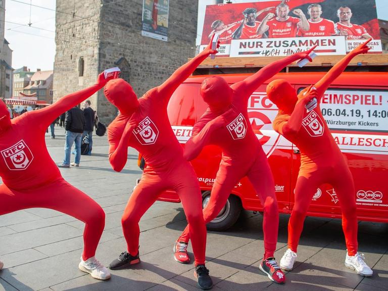 Vier Menschen in Ganzkörperkostümen, die auch das Gesicht verhüllen, stehen vor einem roten VW Bus, der mit Werbung des Halleschen FC bedruckt ist. Sie zeigen in den Himmel.