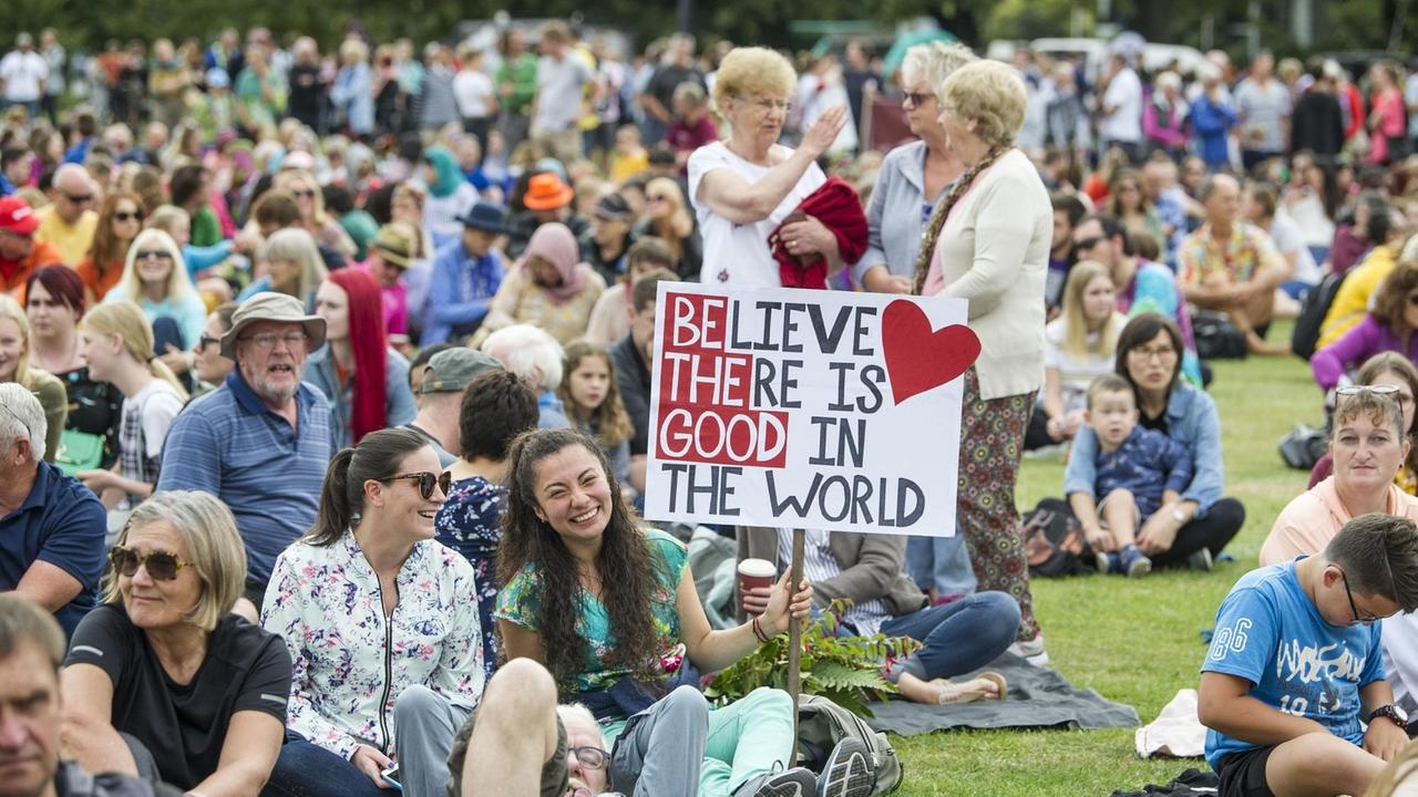 Viele Menschen sitzen fröhlich auf einer grünen Wiese. Eine junge Frau hält ein Schild hoch, auf dem steht: "Glaube an das  Gute in der Welt."